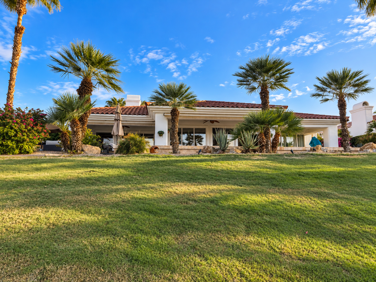 Rear exterior of Solmirage villa viewed from the fairway — white stucco architecture, terracotta roof, manicured lawn, towering palms, agave, and a vivid blue desert sky